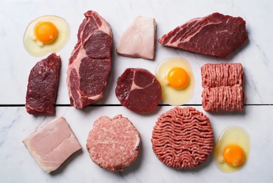 Assortment of raw ribeye steak, beef patties, pork, and fresh eggs arranged on white marble.